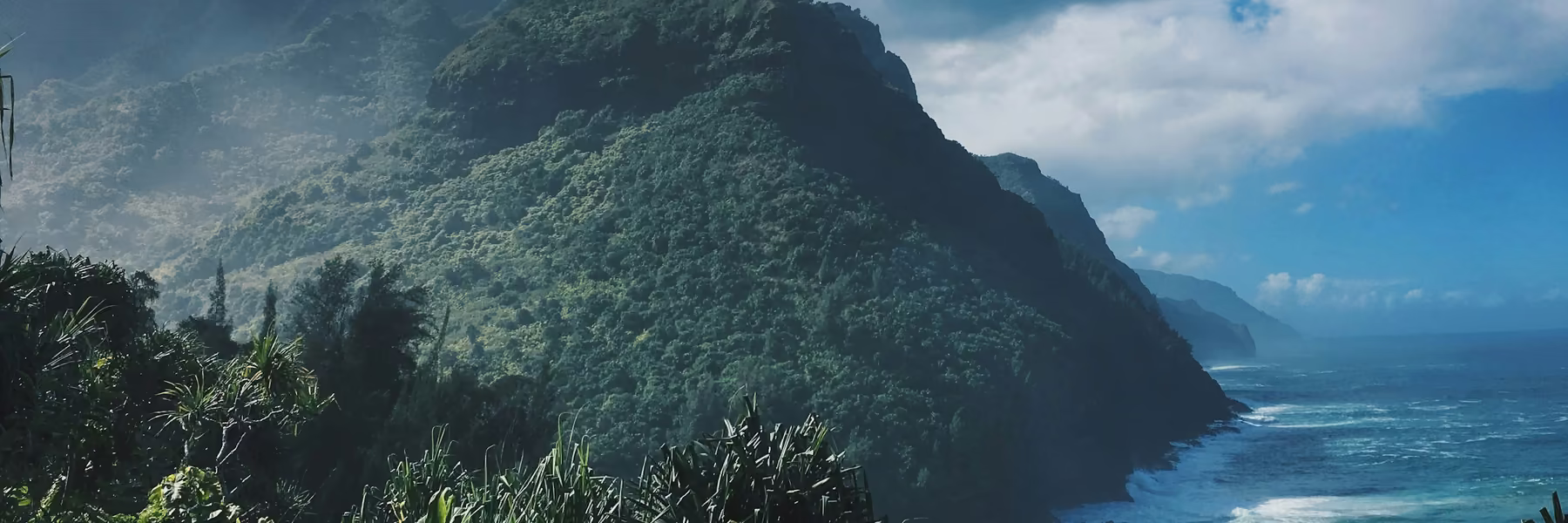 A steep green ridge falling to a turquoise shoreline, with surf breaking along the coast below.