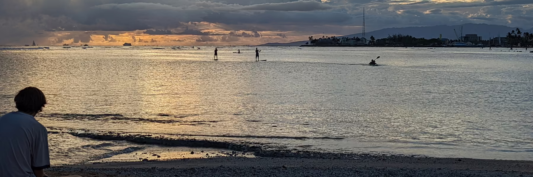 Evening on a calm shoreline, with paddleboarders offshore and a city skyline beyond.