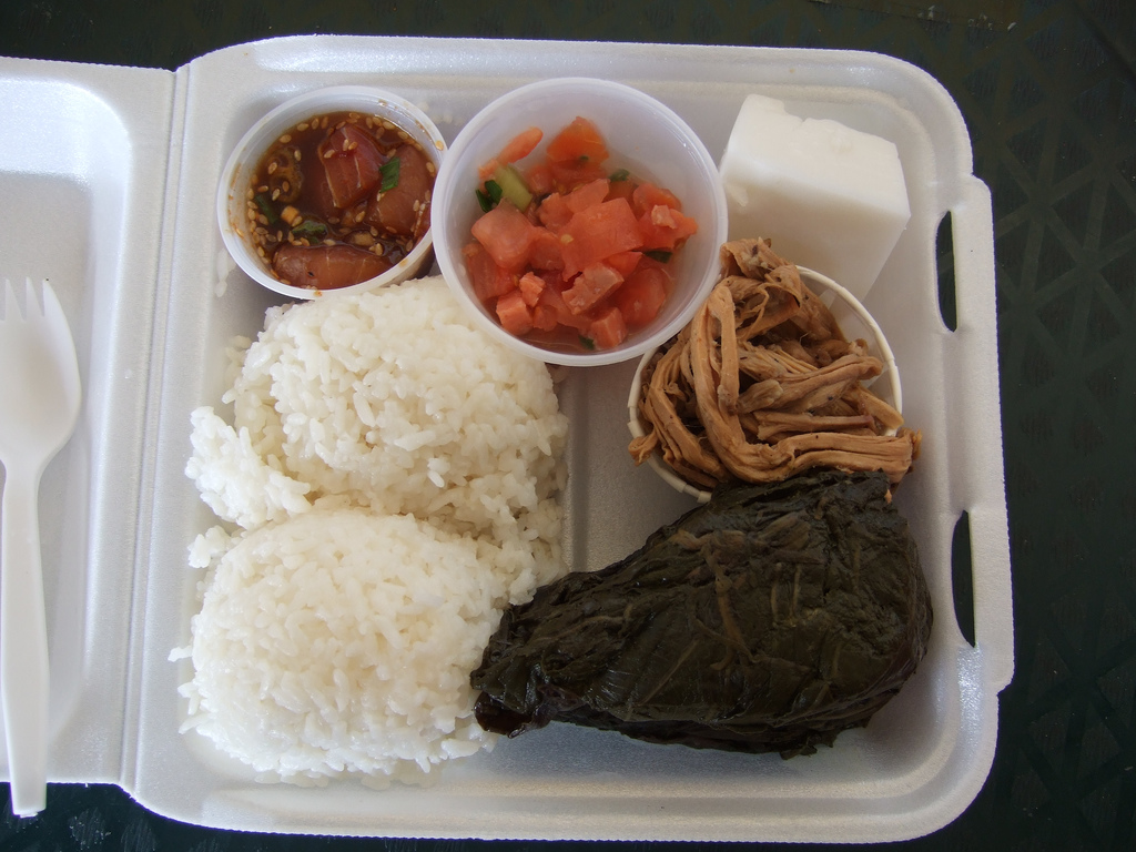Plate lunch in a foam clamshell with rice, poke, lomi salmon, haupia, kalua pork, and lau lau.